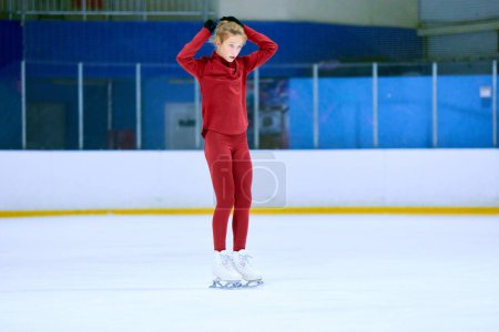 Foto de Chica adolescente, atleta de patinaje artístico en ropa deportiva roja sentirse cansado después de la sesión de entrenamiento duro, la fijación del cabello, de pie en la pista de hielo arena. Concepto de deporte profesional, competición, escuela, pasatiempo, anuncio - Imagen libre de derechos