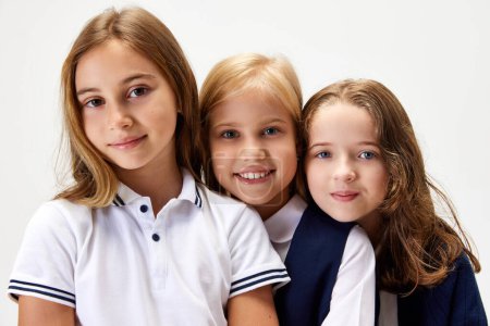 Photo for Three beautiful children, little girls, friends wearing school uniform, standing against white studio background. Concept of childhood, school, education, fashion, style. Copy space for ad - Royalty Free Image