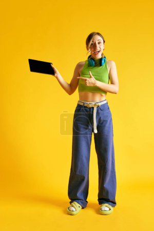 Téléchargez les photos : Image pleine longueur de belle jeune femme en vêtements élégants pointant sur l'écran de la tablette sur fond de studio jaune. Les ventes. Concept d'émotions humaines, jeunesse, mode, mode de vie, publicité - en image libre de droit