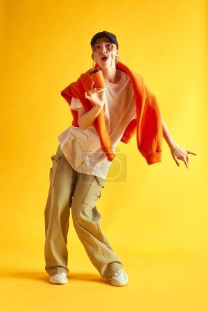 Foto de Imagen de larga duración de una mujer joven con ropa deportiva elegante bailando, divirtiéndose contra el fondo amarillo del estudio. Concepto de emociones humanas, juventud, moda, estilo de vida, anuncio - Imagen libre de derechos