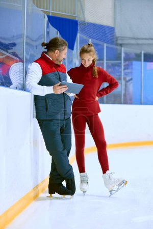 Foto de Chica, atleta de patinaje artístico con entrenador mirando en la tableta, técnicas de aprendizaje, viendo videos en pista de hielo arena. Concepto de deporte profesional, competición, escuela de deporte, salud, hobby, anuncio - Imagen libre de derechos