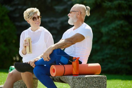 Photo for Cheerful, positive, smiling mature people, man and woman sitting on bench outdoors with fitness mat. After training. Concept of active and sportive lifestyle, age, health care, exercising, ad - Royalty Free Image