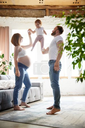 Foto de Padres cariñosos felices, hombre y mujer embarazada jugando con una niña pequeña, hija en casa. Concepto de embarazo, familia, amor, relación, paternidad e infancia - Imagen libre de derechos