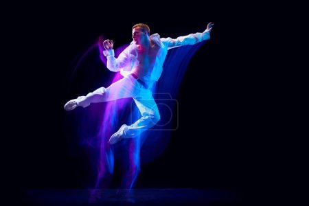 Photo for Dynamic image of stylish young man in white clothes humping over black studio background in neon with mixed lights effect. Concept of movements, art, dance and sport, fashion, youth. Copy space for ad - Royalty Free Image