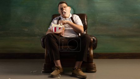 Photo for Creative collage. Fat teen boy wearing retro style clothes sitting in brown armchair on dark vintage background. Watching TV show, eats popcorn. Overweight, carefree, weight loss, health - Royalty Free Image