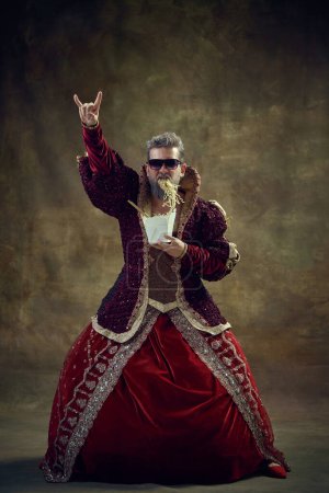 Photo for Gray-haired man in image of medieval royal person, king in female dress eating instant noodles on vintage background. Historical retrospectives, fashion, provoking projects, gender fluidity concept - Royalty Free Image