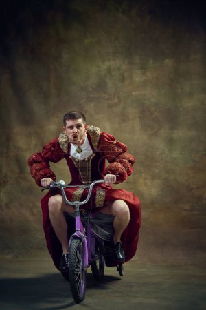Téléchargez les photos : Jeune homme, personne royale médiévale en robe féminine chevauchant petit vélo sur fond vintage. Des émotions humoristiques. Concept de rétrospectives historiques, mode, projets provoquant, fluidité des genres - en image libre de droit