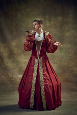 Photo for Young man in image of medieval royal person wearing female dress, attire and talking on mobile phone on vintage background. Concept of historical retrospectives, fashion, provoking projects, gadgets - Royalty Free Image