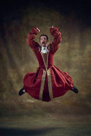 Photo for Young emotional man, medieval royal person emotionally jumping in female dress against vintage background. Concept of historical retrospectives, fashion, provoking projects, gender fluidity - Royalty Free Image
