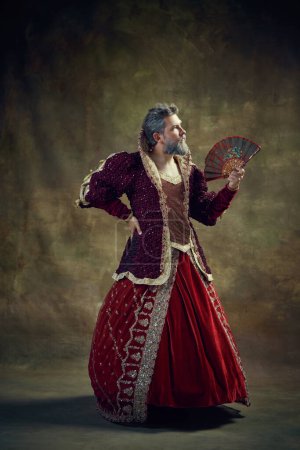 Photo for Monarchs wardrobe. Medieval nobleman, royal person, king in female dress with fan against vintage background. Concept of historical retrospectives, fashion, provoking projects, gender fluidity - Royalty Free Image