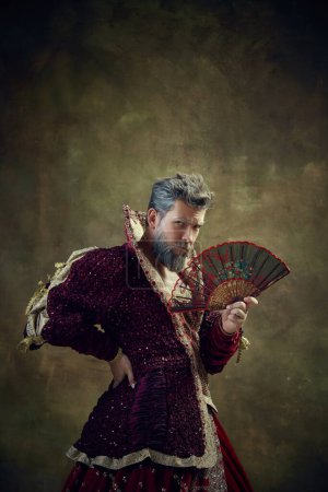 Photo for Monarchs wardrobe. Serious medieval nobleman, royal person, king in female dress with fan over vintage background. Concept of historical retrospectives, fashion, provoking projects, gender fluidity - Royalty Free Image