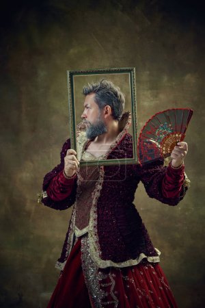 Photo for Portrait of serious nobleman, medieval king wearing female dress with fan against vintage green background. Historical photography. Concept of historical retrospectives, fashion, provoking projects - Royalty Free Image