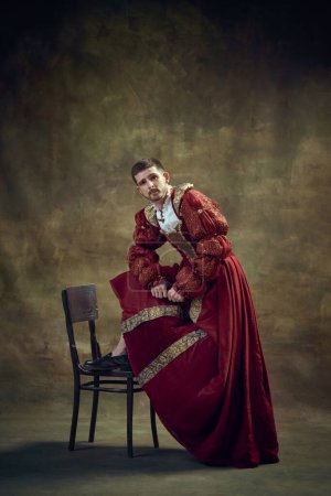Photo for Young medieval prince, royal person leaning on chair, wearing female dress on dark green background. Renaissance style. Historical retrospectives, fashion, provoking projects, gender fluidity concept - Royalty Free Image