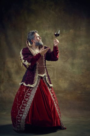 Foto de Rey medieval de pelo gris en vestido femenino levantando copa de vino sobre fondo vintage. Bodega y degustación. Concepto de retrospectivas históricas, moda, proyectos provocadores, fluidez de género - Imagen libre de derechos