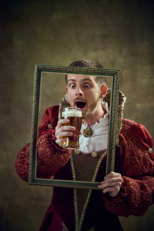 Photo for Medieval royal person, prince in female dress holding picture frame and drinking beer against dark green background. Concept of historical retrospectives, fashion, provoking projects, festival - Royalty Free Image