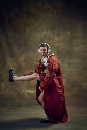 Photo for Young man, medieval prince, monarch in female dress listening to music in headphones, dancing on vintage background. Concept of historical retrospectives, fashion, provoking projects, gender fluidity - Royalty Free Image