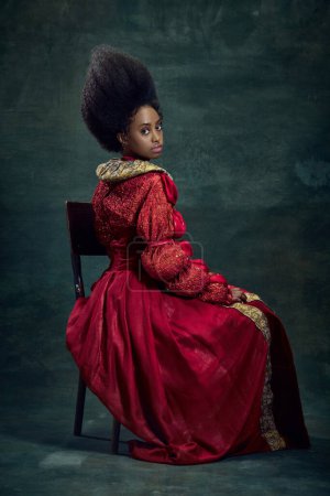 Téléchargez les photos : Portrait de belle, élégante jeune femme africaine, princesse médiévale en robe vintage assise, posant sur fond vert foncé. Concept d'histoire, beauté et mode, comparaison des époques, publicité - en image libre de droit