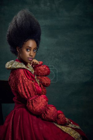 Royal Elegance. Portrait of young, beautiful african woman, medieval princess in vintage dress sitting, posing against dark green background. Concept of history, beauty and fashion, comparison of eras