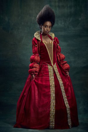 Téléchargez les photos : Costume historique. Portrait de la personne royale médiévale, reine en robe rouge debout sur fond vert vintage. Concept d'histoire, beauté et mode, comparaison des époques, publicité - en image libre de droit