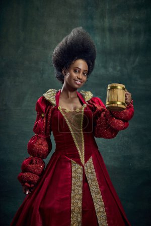 Téléchargez les photos : Positif, souriant jeune femme africaine, reine médiévale en robe rouge élevant tasse avec de la bière sur fond vert vintage. Concept d'histoire, beauté et mode, comparaison des époques, publicité - en image libre de droit