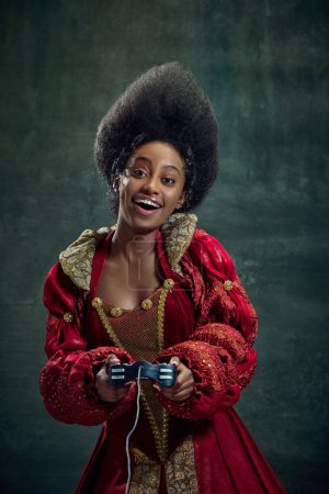 Téléchargez les photos : Heureuse, souriante, jeune femme africaine, princesse en robe rouge élégante jouant à des jeux en ligne avec console sur fond vert vintage. Concept d'histoire, beauté et mode, comparaison des époques, publicité - en image libre de droit