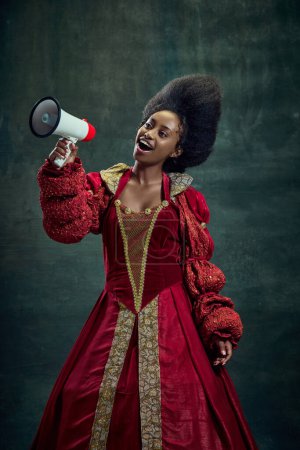 Téléchargez les photos : Jeune femme africaine positive, noble médiévale, reine en robe rouge parlant en mégaphone sur fond vert vintage. Concept d'histoire, beauté et mode, comparaison des époques, ventes, nouvelles, publicité - en image libre de droit