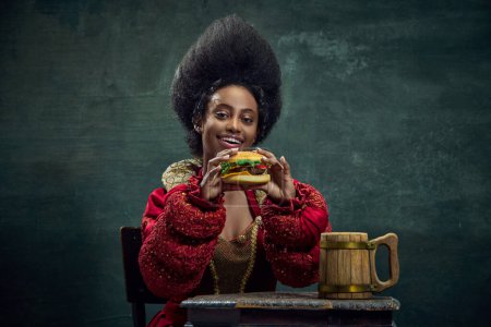 Téléchargez les photos : Belle femme africaine, princesse médiévale mangeant un délicieux hamburger sur fond vert vintage. Restauration rapide. Concept d'histoire, beauté et mode, comparaison des époques, publicité - en image libre de droit