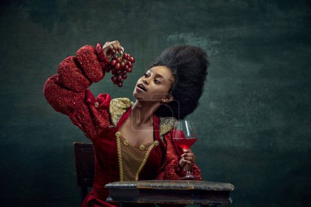 Téléchargez les photos : Une cave. Jeune femme africaine, princesse, reine en robe rouge élégante mangeant des raisins et buvant du vin sur fond vert vintage. Concept d'histoire, beauté et mode, comparaison des époques, publicité - en image libre de droit