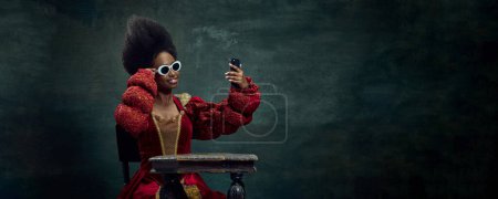 Téléchargez les photos : Jeune femme africaine, reine, princesse en robe rouge et lunettes de soleil prenant selfie avec téléphone portable sur fond vintage. Concept d'histoire, beauté et mode, comparaison des époques, médias sociaux, publicité - en image libre de droit
