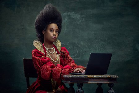 Téléchargez les photos : Princesse médiévale, jeune femme africaine en élégant costume rouge travaillant sur ordinateur portable sur fond vert vintage. Concept d'histoire, de beauté et de mode, comparaison des époques, des affaires, de la publicité - en image libre de droit