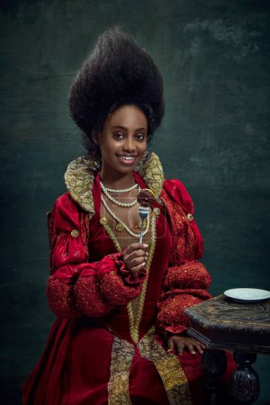 Téléchargez les photos : Jeune femme africaine, reine médiévale, princesse en robe rouge mangeant des bonbons, avec des petits beignets sur fond vert vintage. Concept d'histoire, beauté et mode, comparaison des époques, publicité - en image libre de droit