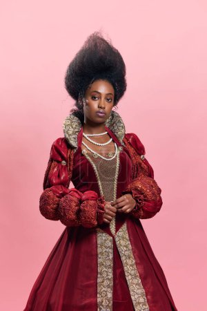 Téléchargez les photos : Portrait de belle jeune femme africaine, princesse médiévale en robe vintage posant sur fond de studio rose. Fierté culturelle. Concept d'histoire, beauté et mode, comparaison des époques, publicité - en image libre de droit