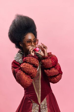 Foto de Mujer africana con estilo en la imagen de la persona real medieval, con vestido rojo y gafas de sol, que sopla globo sobre fondo de estudio rosa. Concepto de historia, belleza y moda, comparación de épocas, ad - Imagen libre de derechos