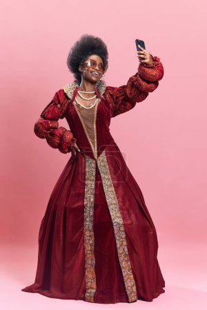Téléchargez les photos : Jeune femme africaine, reine, princesse en robe rouge et lunettes de soleil prenant selfie avec téléphone portable sur fond de studio rose. Influenceur. Concept d'histoire, beauté et mode, comparaison des époques, publicité - en image libre de droit