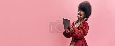Téléchargez les photos : Princesse médiévale, jeune femme africaine en robe rouge élégante à l'aide d'une tablette sur fond vert vintage. Shopping et loisirs en ligne. Concept d'histoire, beauté et mode, comparaison des époques, publicité - en image libre de droit