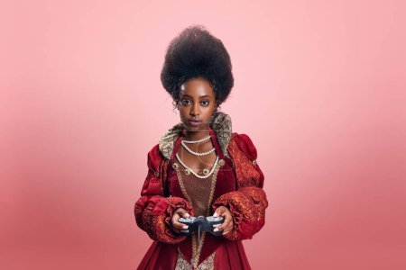 Téléchargez les photos : Jeune femme africaine, princesse en robe rouge élégante jouant à des jeux en ligne avec console contre fond de studio rose. Concept d'histoire, beauté et mode, comparaison des époques, publicité - en image libre de droit