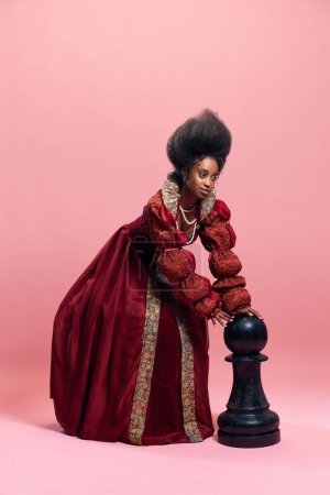 Photo for Checkmate. Portrait of medieval princess, queen in red costume playing with giant chess piece against pink studio background. Concept of history, beauty and fashion, comparison of eras, ad - Royalty Free Image