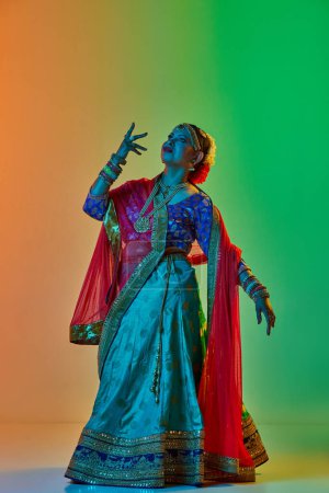 Foto de Elegante, madura, hermosa mujer india en ropa tradicional, vestido posando contra el fondo del estudio degradado en luz de neón. Concepto de belleza, moda, India, tradiciones, coreografía, arte. Anuncio - Imagen libre de derechos