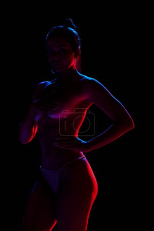Photo for Sensual, beautiful woman posing in underwear, covering breast with hand against black studio background in neon lights. Concept of female beauty, body art, aesthetics, femininity and sensuality. - Royalty Free Image