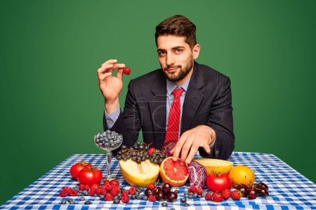 Photo for Man in a suit sitting at table against green background and eating fresh berries, fruits and vegetables. Concept of food, creativity, organic products, health. Pop art photography. Copy space for ad - Royalty Free Image