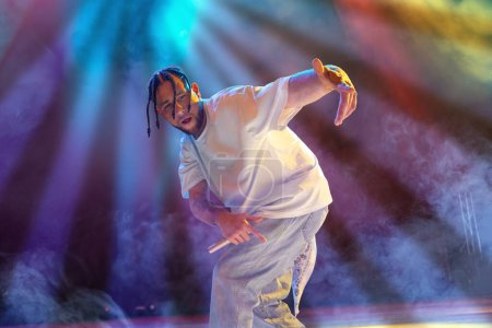 Photo for Rap musician. Young artistic man with dreads performing on stage over multicolored neon lights. Concert at night club. Concept of music, performance, art, talent, nightlife, joy, party, lifestyle, ad - Royalty Free Image