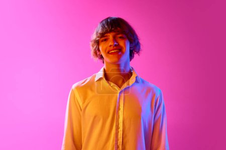 Foto de Retrato de un joven con camisa blanca mirando a la cámara y sonriendo sobre el fondo rosa del estudio con luces de neón. Concepto de emociones humanas, expresión facial, juventud, estilo de vida. Anuncio - Imagen libre de derechos