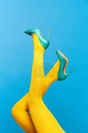 Foto de Piernas delgadas femeninas que usan zapatos elegantes, con tacón, verdes y medias amarillas sobre fondo azul. Fotografía colorida. Concepto de moda, creatividad, imaginación. Copiar espacio para anuncio - Imagen libre de derechos