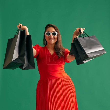 Téléchargez les photos : Belle femme en robe rouge élégante et lunettes de soleil debout avec de nombreux sacs à provisions sur fond de studio vert. Les ventes. Vendredi noir. Concept de beauté, émotions humaines, style de vie, mode, publicité - en image libre de droit