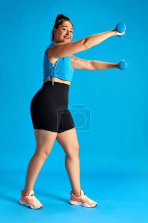 Foto de Joven mujer sonriente con sobrepeso en entrenamiento de ropa deportiva, haciendo ejercicios con mancuernas sobre fondo azul. Perder peso. Concepto de deporte, estilo de vida saludable, emociones humanas, moda, anuncio - Imagen libre de derechos