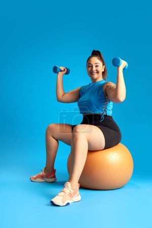 Foto de Mujer positiva con sobrepeso en ropa deportiva, sentada en una pelota de fitness y entrenando con pesas sobre fondo azul. Concepto de deporte, estilo de vida saludable, emociones humanas, moda, anuncio - Imagen libre de derechos