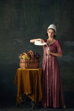 Téléchargez les photos : Elégante jeune femme en robe Renaissance, bonne versant du lait dans du verre sur fond vintage sombre. Une paysanne. Concept d'histoire, comparaison des époques, beauté, art, créativité - en image libre de droit
