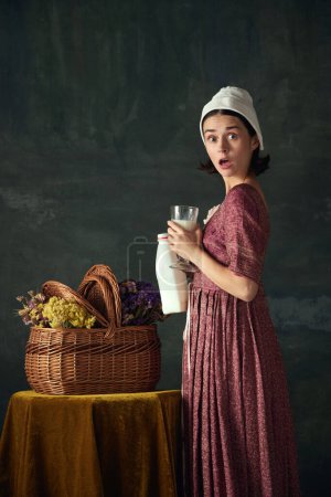 Photo for Shock and unexpected meeting. Emotive young woman, medieval maid with standing with milk against dark vintage background. Concept of history, comparison of eras, beauty, art, creativity - Royalty Free Image