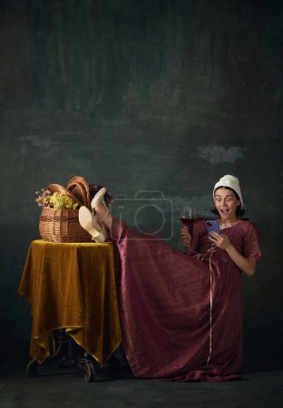 Foto de Mujer joven emocionada en la imagen de la persona medieval, criada mirando en el teléfono móvil y beber vino sobre fondo verde vintage. Concepto de historia, comparación de épocas, belleza, arte, creatividad - Imagen libre de derechos