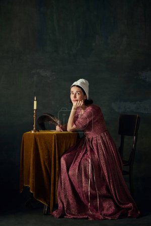 Foto de Mujer joven bonita y soñadora en la imagen de una doncella renacentista sentada a la mesa y escribiendo una carta sobre un oscuro fondo vintage. Concepto de historia, comparación de épocas, belleza, arte, creatividad - Imagen libre de derechos
